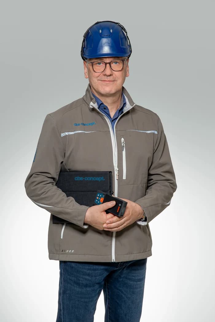 Maik Taute von cbs-concept in Arbeitsjacke mit blauem Schutzhelm, Projektmappe und Messgerät in der Hand
